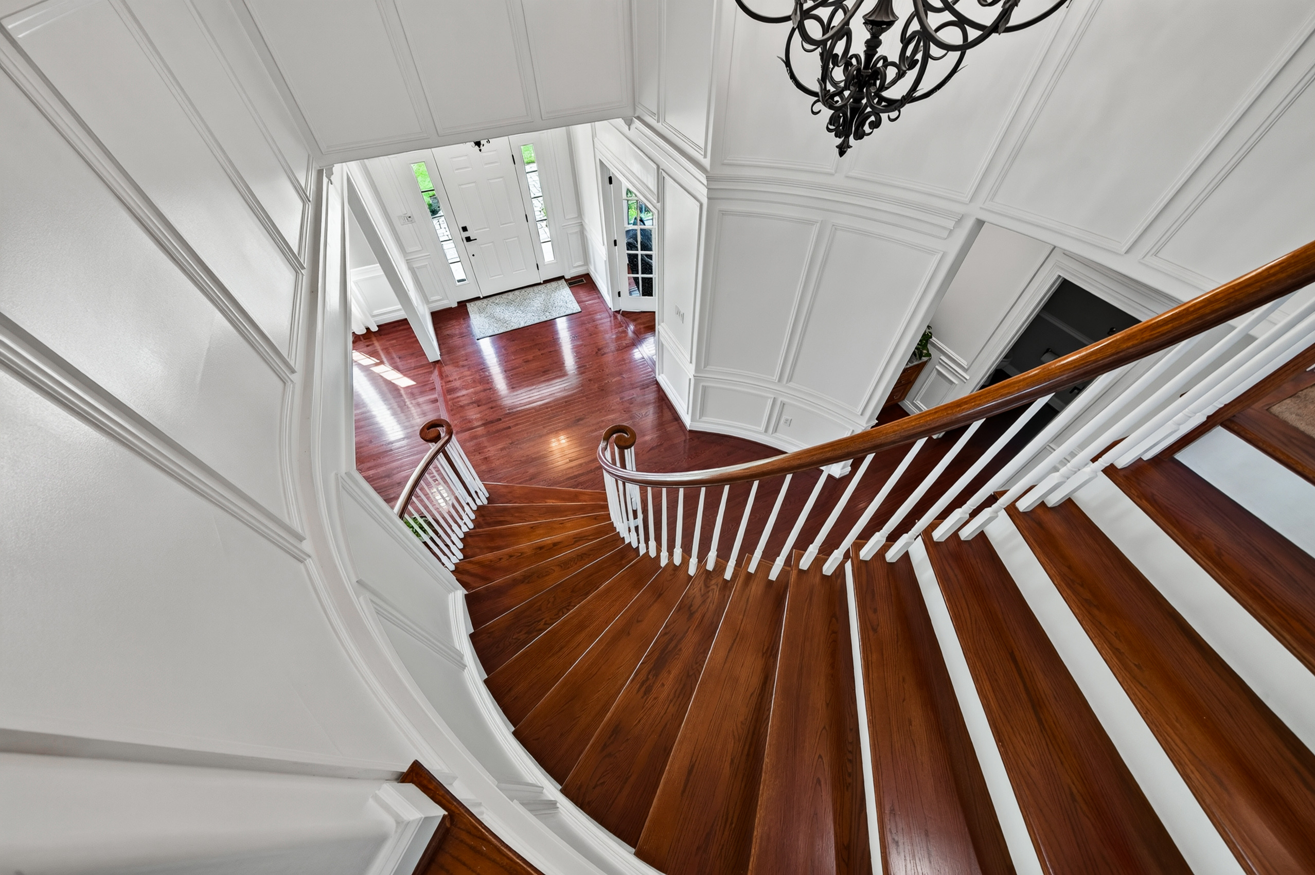 Curved staircase descending into the foyer beneath two stories of paneled wainscoting and a wrought-iron chandelier